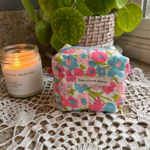 May include: A small, square floral print pouch with pink and blue flowers on a white background. The pouch has a zipper closure and a small label that reads "Moon Altered Atelier". A lit candle and a potted plant are in the background.