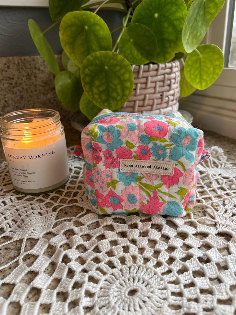 May include: A small, square floral print pouch with pink and blue flowers on a white background. The pouch has a zipper closure and a small label that reads "Moon Altered Atelier". A lit candle and a potted plant are in the background.