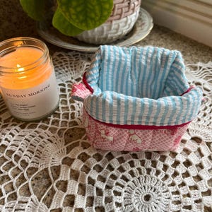May include: A small, handmade fabric basket with a pink and white dog pattern exterior and a blue and white striped interior. The rim is trimmed with red fabric. A lit candle in a glass jar is visible in the background.