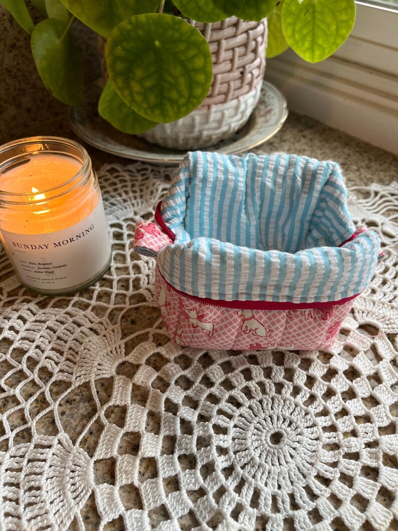 May include: A small, handmade fabric basket with a pink and white dog pattern exterior and a blue and white striped interior. The rim is trimmed with red fabric. A lit candle in a glass jar is visible in the background.