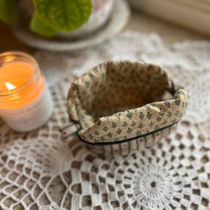 May include: A small, open fabric basket with a beige interior and a patterned exterior featuring small green floral designs. The basket has a dark green trim and is set on a white lace doily. A lit candle and a potted plant are in the background.