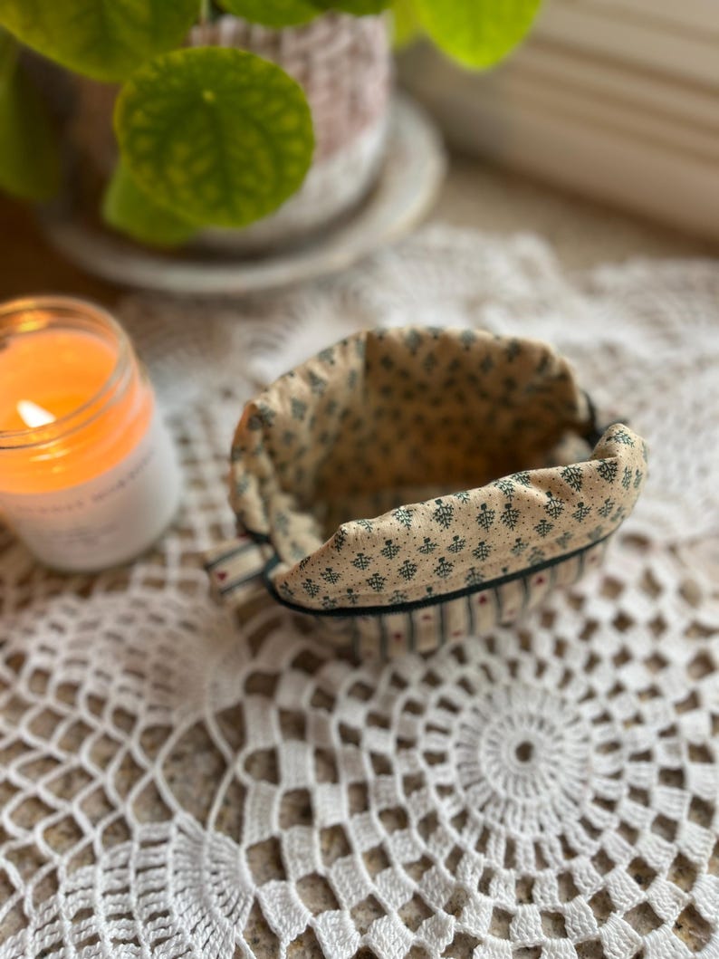 May include: A small, open fabric basket with a beige interior and a patterned exterior featuring small green floral designs. The basket has a dark green trim and is set on a white lace doily. A lit candle and a potted plant are in the background.