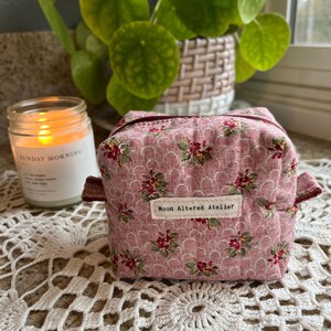May include: A small, pink floral patterned fabric box with a zipper closure and a label that reads "Moon Altered Atelier." The box has a quilted design and is accented with burgundy trim. A lit candle and a potted plant are in the background.