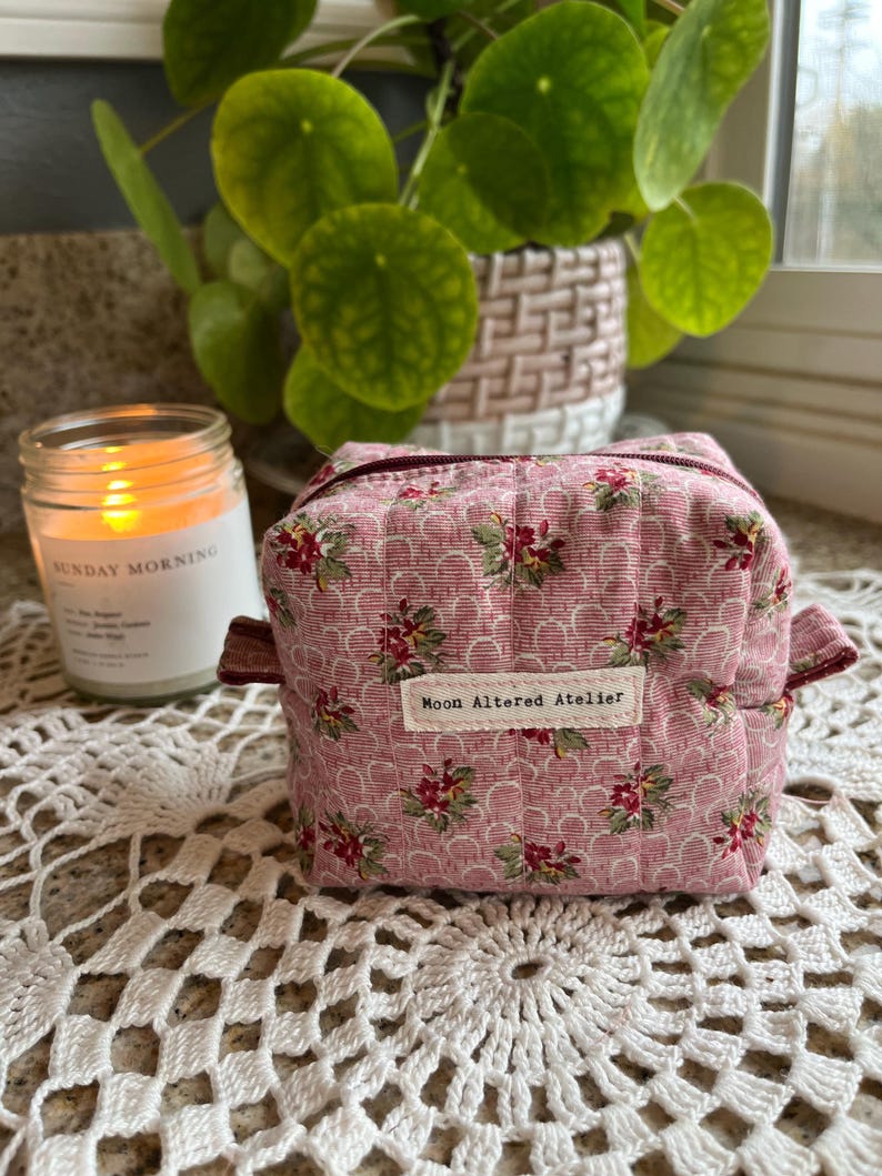 May include: A small, pink floral patterned fabric box with a zipper closure and a label that reads "Moon Altered Atelier." The box has a quilted design and is accented with burgundy trim. A lit candle and a potted plant are in the background.
