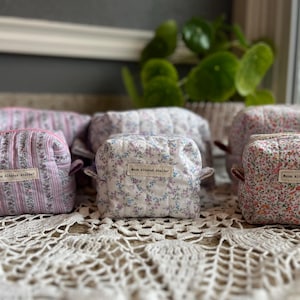 May include: A collection of small, quilted cosmetic bags in floral and striped designs. The bags feature pink, white, and lavender hues. Each bag is labelled with "Moon Altered Atelier".
