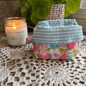 May include: A small, fabric storage basket with a floral pattern and a blue and white striped top. The basket sits on a white crocheted doily, with a lit candle in the background. The candle has the text "SUNDAY MORNING" on the label.