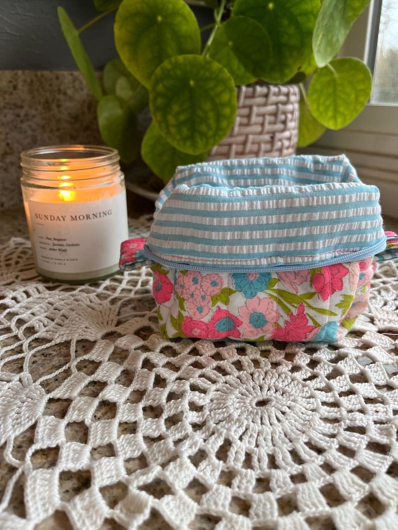 May include: A small, fabric storage basket with a floral pattern and a blue and white striped top. The basket sits on a white crocheted doily, with a lit candle in the background. The candle has the text "SUNDAY MORNING" on the label.