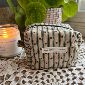 May include: A small, rectangular beige cosmetic bag with a striped pattern and small red hearts. The bag has a zipper closure and a label that reads "Moon Altered Atelier". The bag is sitting on a white lace surface.