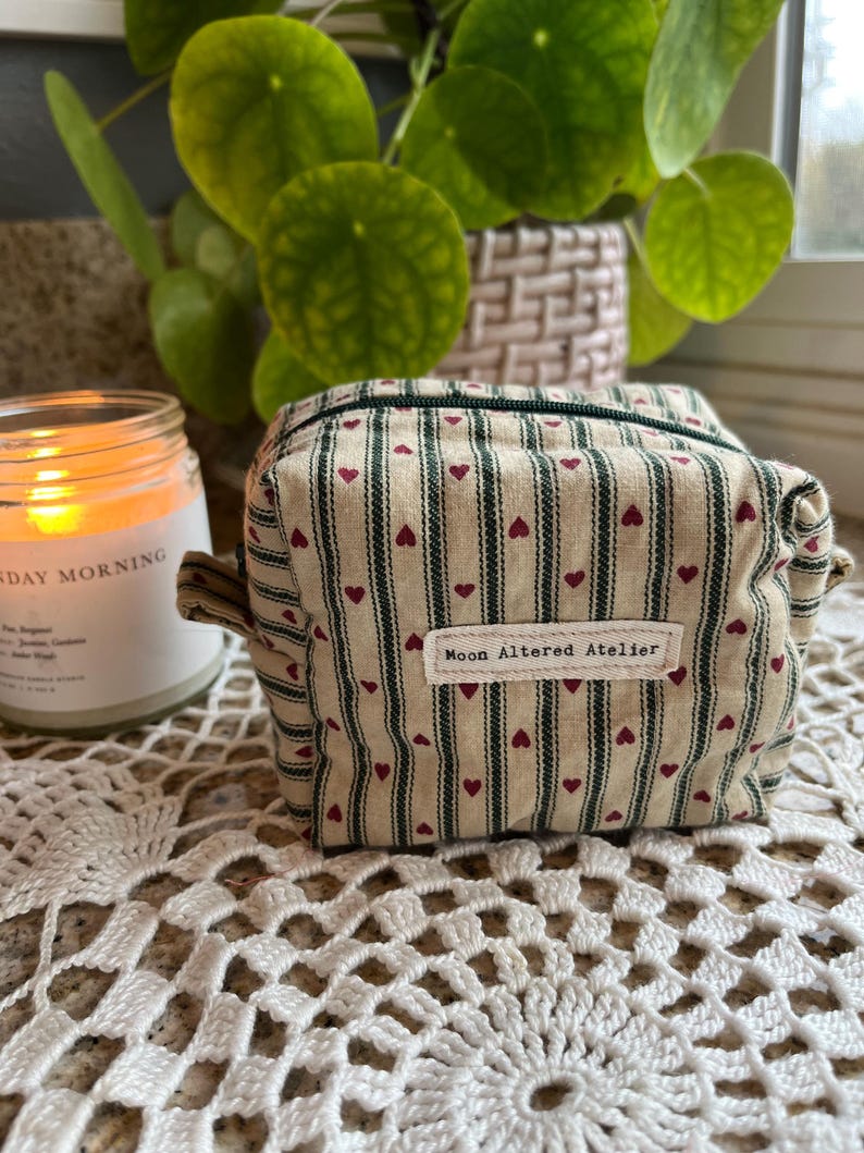 May include: A small, rectangular beige cosmetic bag with a striped pattern and small red hearts. The bag has a zipper closure and a label that reads "Moon Altered Atelier". The bag is sitting on a white lace surface.