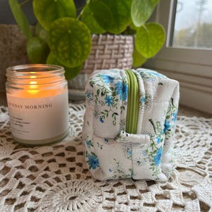 May include: A small, square, white fabric pouch with a blue floral pattern and a green zipper. A lit candle in a glass jar with the text "SUNDAY MORNING" is to the left. A green plant is in the background.