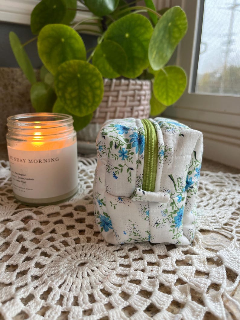 May include: A small, square, white fabric pouch with a blue floral pattern and a green zipper. A lit candle in a glass jar with the text "SUNDAY MORNING" is to the left. A green plant is in the background.