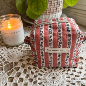 May include: A small, square, zippered pouch with a red and white striped pattern, accented with blue floral designs. The text "Moon Altered Atelier" is displayed on a small label. A lit candle and a plant are in the background.