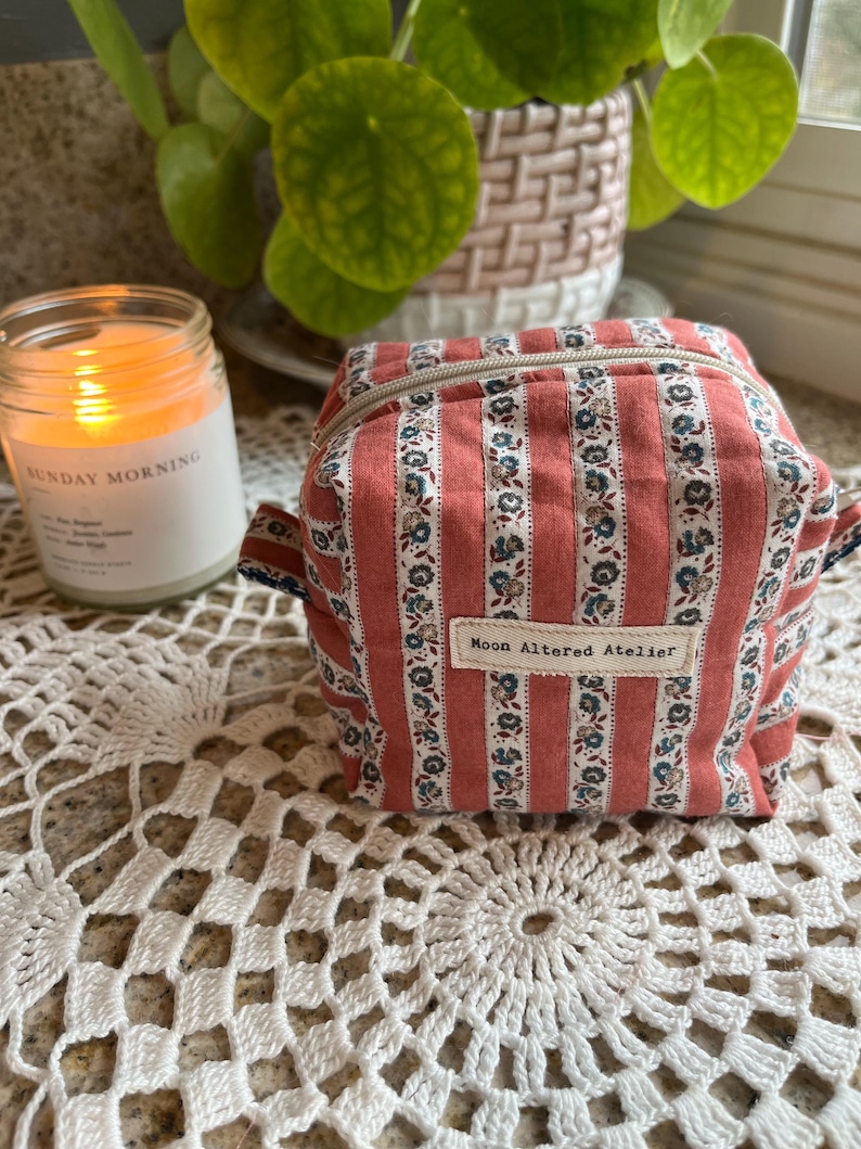 May include: A small, square, zippered pouch with a red and white striped pattern, accented with blue floral designs. The text "Moon Altered Atelier" is displayed on a small label. A lit candle and a plant are in the background.