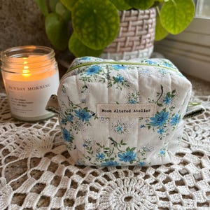 May include: A small, quilted cosmetic bag with a white background and a blue floral pattern. The bag has a green zipper and handles. The text "Moon Altered Atelier" is on a small tag. A lit candle and a potted plant are in the background.