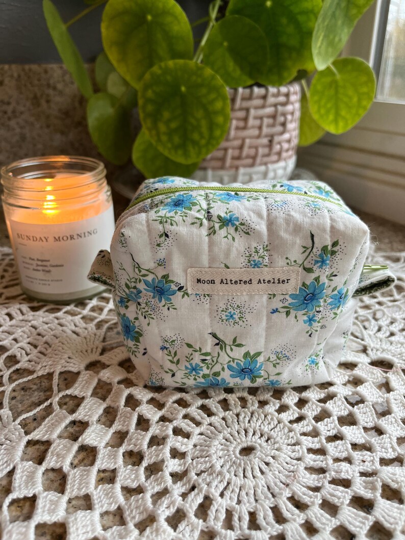 May include: A small, quilted cosmetic bag with a white background and a blue floral pattern. The bag has a green zipper and handles. The text "Moon Altered Atelier" is on a small tag. A lit candle and a potted plant are in the background.