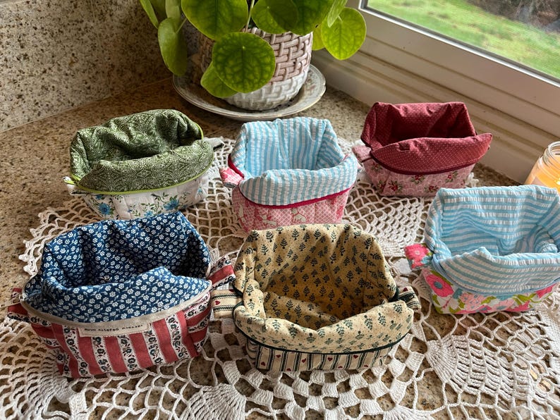 May include: A collection of six fabric baskets in various patterns and colors. The baskets feature floral, striped, and patterned designs in shades of blue, red, green, and yellow. Each basket has a contrasting interior lining.