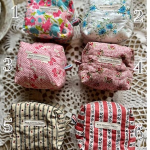 May include: Six small, quilted fabric pouches with floral and striped patterns. Each pouch has a small loop and a label that reads "Moon Altered Atelier". The pouches are in shades of pink, blue, red, and cream.