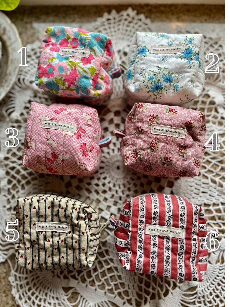 May include: Six small, quilted fabric pouches with various floral and striped patterns. Each pouch has a small loop and a label that reads "Moon Altered Atelier". The pouches are in shades of pink, blue, red, and cream.