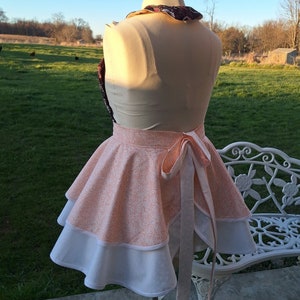 May include: A pink and white apron with a ruffled skirt and a bow tie. The apron is made of a cotton fabric with a swirl pattern.
