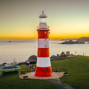 Puede incluir: Un faro rojo y blanco a rayas con una linterna de vidrio en la parte superior, de pie en una colina cubierta de hierba con vistas a una masa de agua al atardecer. El faro está rodeado por una pasarela circular con luces.