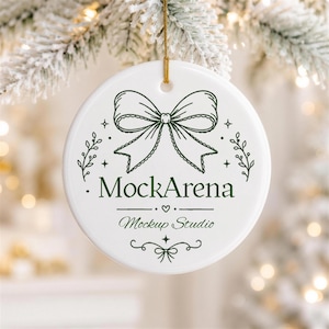 May include: A round white ceramic ornament featuring a dark green line art illustration of a bow at the top. The text MockArena is printed in the centre, followed by the words Mockup Studio below it, accented by small stars and flourishes.