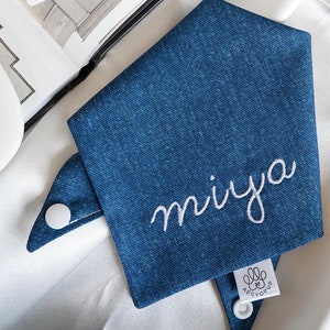 Pode incluir: Um bandana de brim azul com costuras brancas e o nome "miya" bordado em branco. O bandana tem um botão branco em cada extremidade e uma etiqueta branca com o texto "AEGYOPUP" e uma pegada.