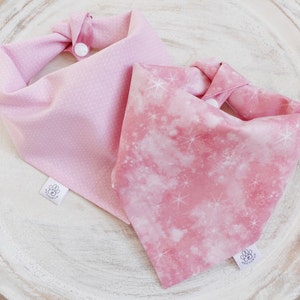 May include: Two pink dog bandanas with white polka dots and a pink and white galaxy print. Both bandanas have a white snap closure and a white label with a paw print logo.