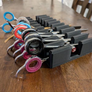 May include: A collection of professional hair shears with colorful finger rings, including blue, red, pink, and black. The shears are stored in a black organizer on a wooden surface. The blades are silver and the handles have various colored inserts.