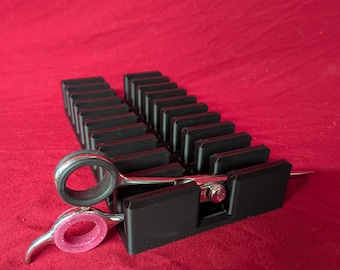 May include: A black plastic storage rack with multiple vertical slots designed to hold tools, featuring a pair of silver metal grooming shears with pink and black rubber finger inserts resting horizontally across the front of the organizer.