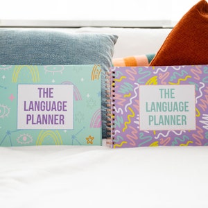 May include: Two colourful language planner notebooks with a spiral binding. The notebook on the left has a light blue cover with a rainbow, stars, and eyes design. The notebook on the right has a purple cover with a colourful abstract design. Both notebooks have the text "The Language Planner" on the front.