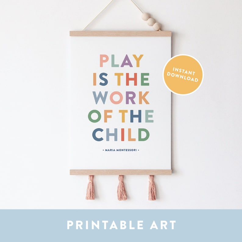 May include: Printable wall art with the quote "Play is the work of the child" by Maria Montessori. The quote is written in colorful letters on a white background. The art is framed with a wooden frame and has pink tassels hanging from the bottom.
