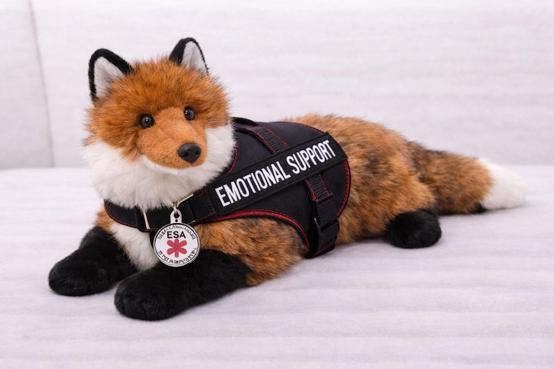 May include: A plush fox toy with brown and white fur, wearing a black vest that reads "EMOTIONAL SUPPORT." The vest has red stitching and a silver ESA tag. The fox is lying down.