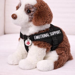 May include: A plush dog with brown and white fur, wearing a black vest that reads "EMOTIONAL SUPPORT." The dog has a small tag with text and a red cross. The dog is sitting on a white surface.