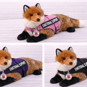 May include: A plush fox toy with brown and white fur, wearing a vest that says "EMOTIONAL SUPPORT." The vest is available in pink, purple, and black. Each fox has a silver ESA tag.