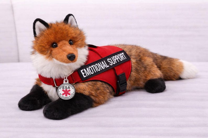 May include: A plush fox toy with brown and white fur, black paws, and a red vest. The vest reads "EMOTIONAL SUPPORT" in white text and has a small ESA tag. The fox is lying down on a light-colored surface.