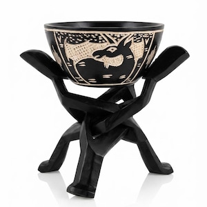 May include: A black carved wooden bowl featuring a light-colored etched design of a goat, resting on a black interlocking wooden stand. The bowl has a smooth finish and the stand is composed of two pieces that cross to support the bowl securely.