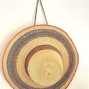 May include: A woven straw hat with a wide brim, featuring concentric circles of natural beige, dark blue, and orange hues. The hat has a red border and is suspended by a brown leather cord, showcasing a rustic, handcrafted aesthetic.