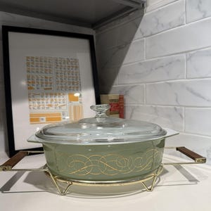 May include: A green casserole dish with a gold swirl design and a glass lid. The dish is sitting on a gold metal stand with wooden handles.