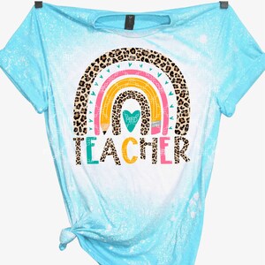 May include: A light blue t-shirt with a bleach-splattered design. The shirt features a rainbow with a leopard print pattern and the word "TEACHER" in colorful letters.
