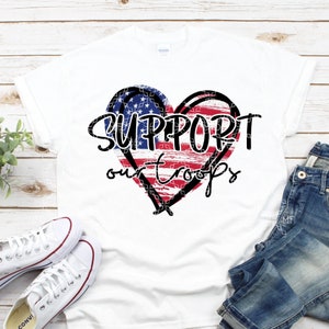 Könnte beinhalten: Weißes T-Shirt mit einem rot-weiß-blauen herzförmigen Design der amerikanischen Flagge. Der Text "SUPPORT our TROOPS" ist in Schwarz über dem Herzen gedruckt.