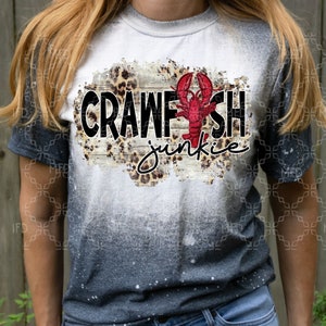 May include: A white t-shirt with a distressed, bleached look. The shirt features a graphic of a red crawfish with the text "Crawfish junkie" in black letters on a woodgrain background with a leopard print overlay.