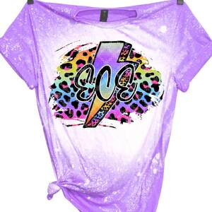 May include: A purple t-shirt with a bleached effect and a rainbow lightning bolt graphic with the word "80s" inside. The graphic is on a leopard print background.