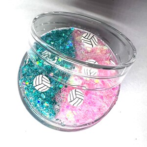 May include: A clear glass jar with a teal and pink glitter mixture and white volleyball charms. The jar has a lid.