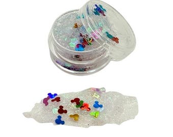 May include: Clear plastic container filled with glitter and colorful Mickey Mouse-shaped confetti. A small pile of glitter and confetti is next to the container. The confetti is in shades of blue, red, yellow, and green.