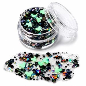 May include: A clear plastic jar filled with a mixture of black, iridescent, and light green mouse-shaped glitter. A small pile of the same glitter mixture is spread out in front of the open jar, showcasing the varied shapes and reflective textures.
