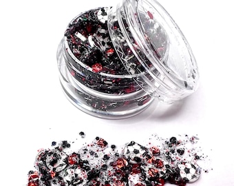 May include: A clear plastic jar filled with black, red, and white glitter. The glitter is shaped like soccer balls. The jar is open and some of the glitter is spilled out on a white surface.