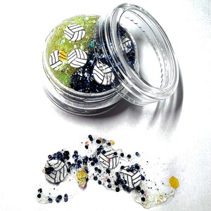 May include: A clear plastic jar filled with green and blue glitter and white volleyball charms. The jar is open and some of the charms have spilled out onto a white surface.