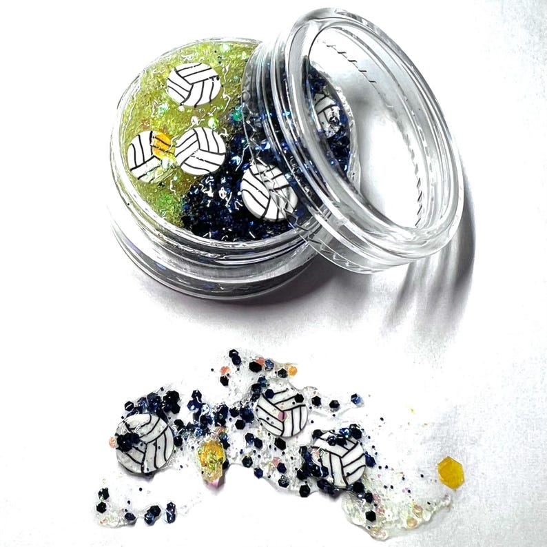 May include: A clear plastic jar filled with green and blue glitter and white volleyball charms. The jar is open and some of the charms have spilled out onto a white surface.