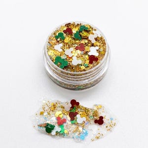 May include: A small clear plastic jar filled with gold glitter and small red, white, and green mouse-shaped sequins. A small pile of the same glitter and mouse-shaped sequins is spread out on the surface directly below the open jar.