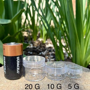 Op de afbeelding: Drie transparante plastic containers met deksels, gelabeld 20 G, 10 G en 5 G. Een Duracell batterij staat naast de containers voor groottevergelijking.